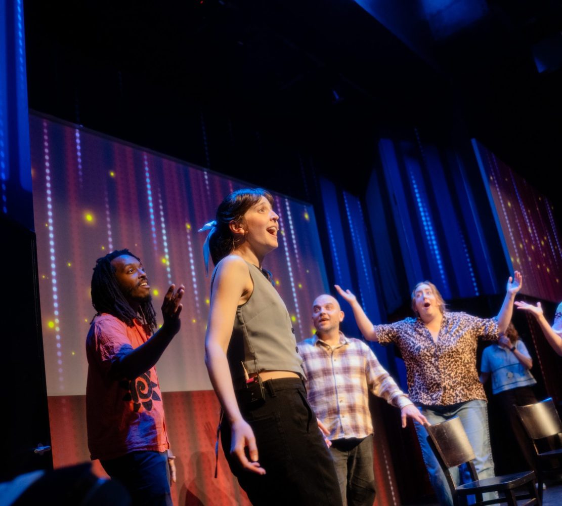 Four people perform on stage with colorful backdrop and chairs.