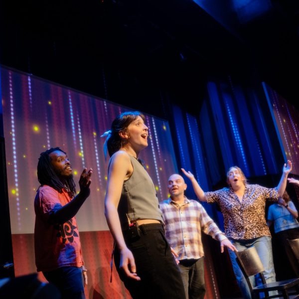 Four people perform on stage with colorful backdrop and chairs.