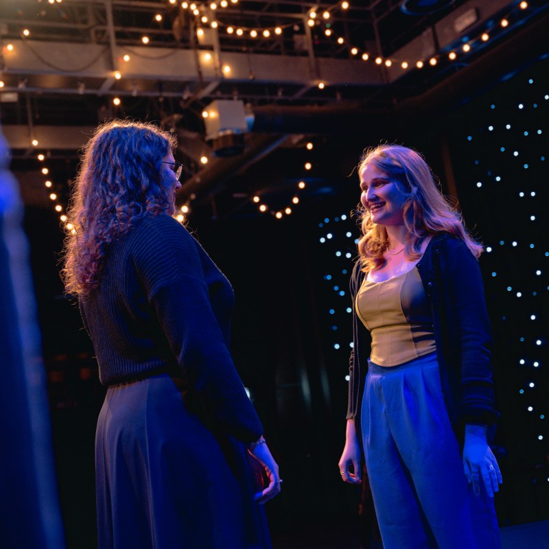 Two women talking on stage under string lights with a starry backdrop.