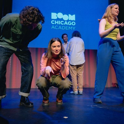 Performers on a stage in front of a Boom Chicago Academy sign, with one crouched and others gesturing.