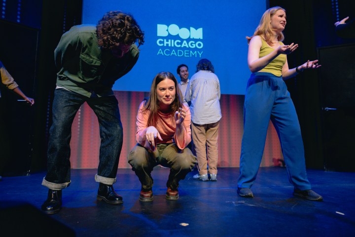 Performers on a stage in front of a Boom Chicago Academy sign, with one crouched and others gesturing.