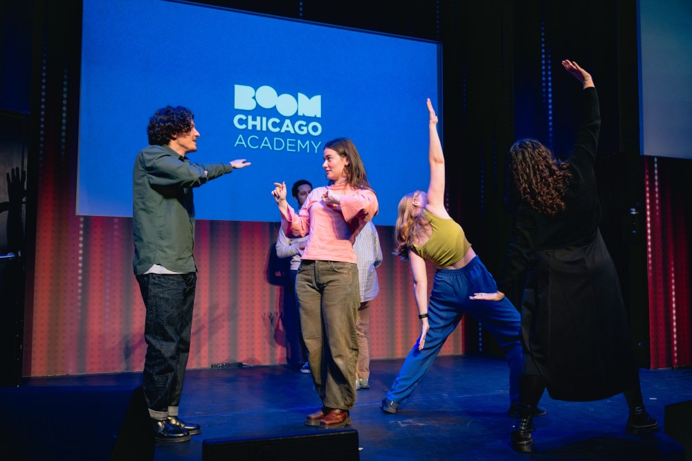 People engaged in improv performance on stage with Boom Chicago Academy sign.