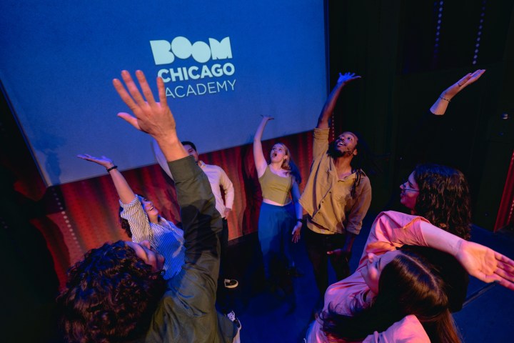 Group of people on stage with arms raised, under 'Boom Chicago Academy' text.