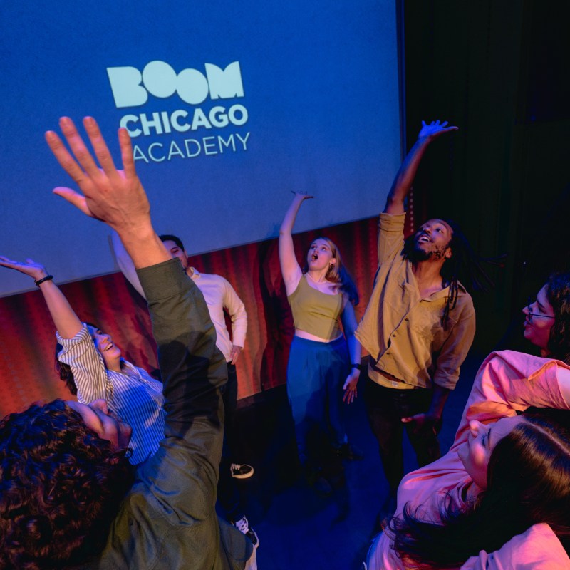 Group of people on stage with arms raised, under 'Boom Chicago Academy' text.