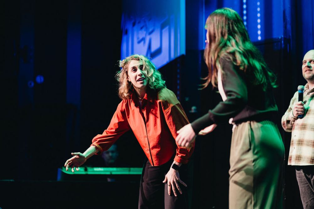 Three people on stage, one in a red shirt gesturing passionately, others watching, with stage lighting.