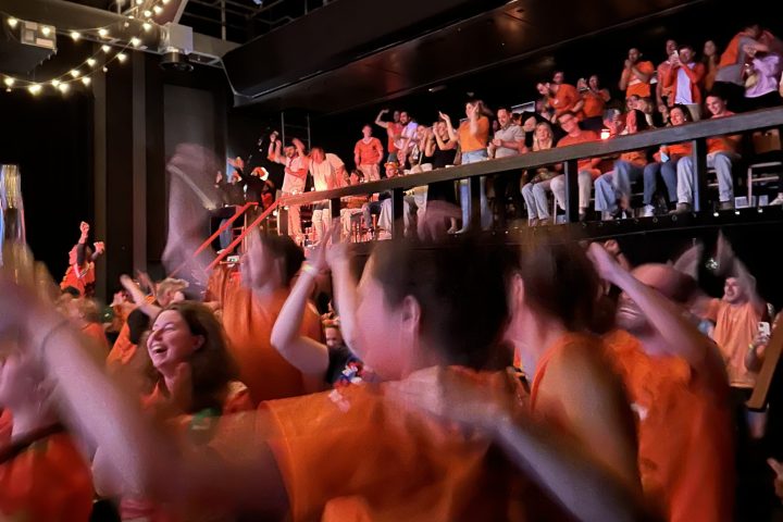 Crowd in orange shirts cheering indoors with string lights overhead.