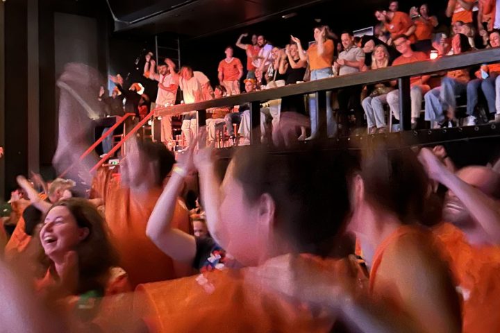 Crowd in orange shirts cheering indoors with string lights overhead.