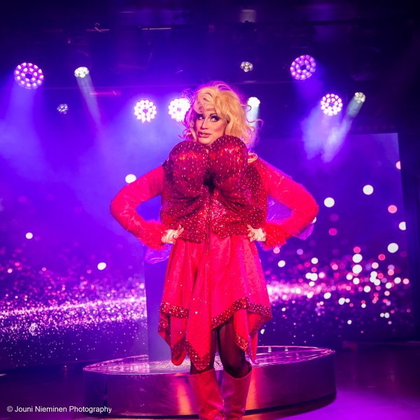Performer in red sequin outfit poses on stage with colorful lights.
