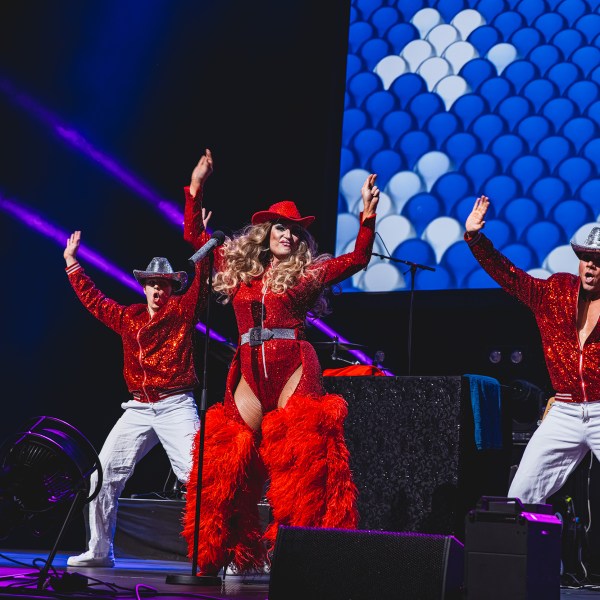 Performer in red outfit and hat dances on stage with two backup dancers in cowboy hats.