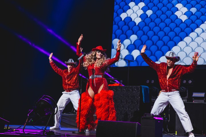 Performer in red outfit and hat dances on stage with two backup dancers in cowboy hats.