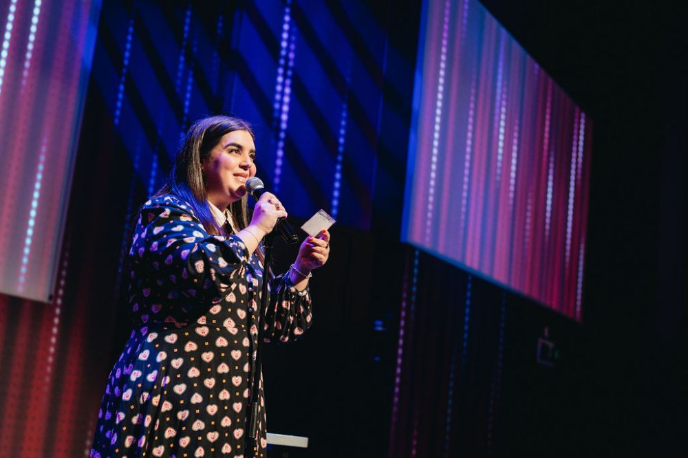 Person in patterned dress speaks into microphone on stage with colorful background.