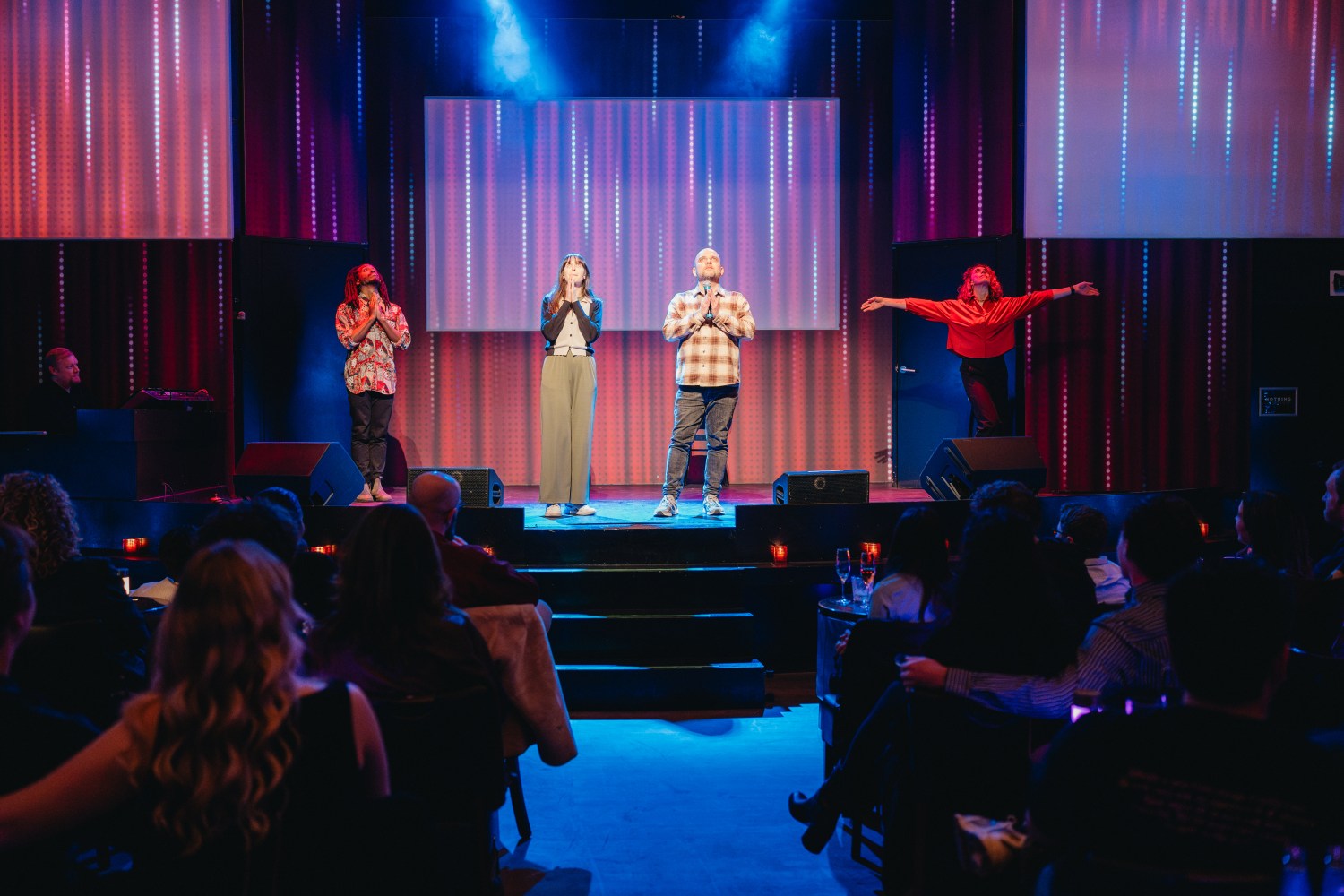 Four performers on stage with colorful lights, audience seated watching.