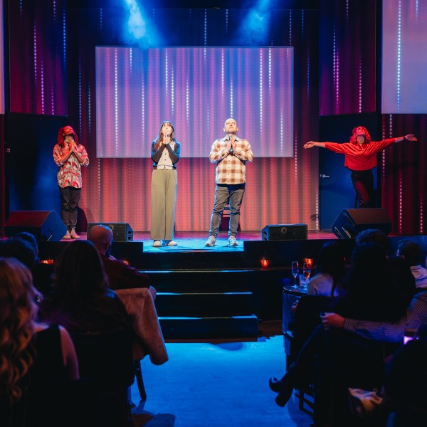 Four performers on stage with colorful lights, audience seated watching.