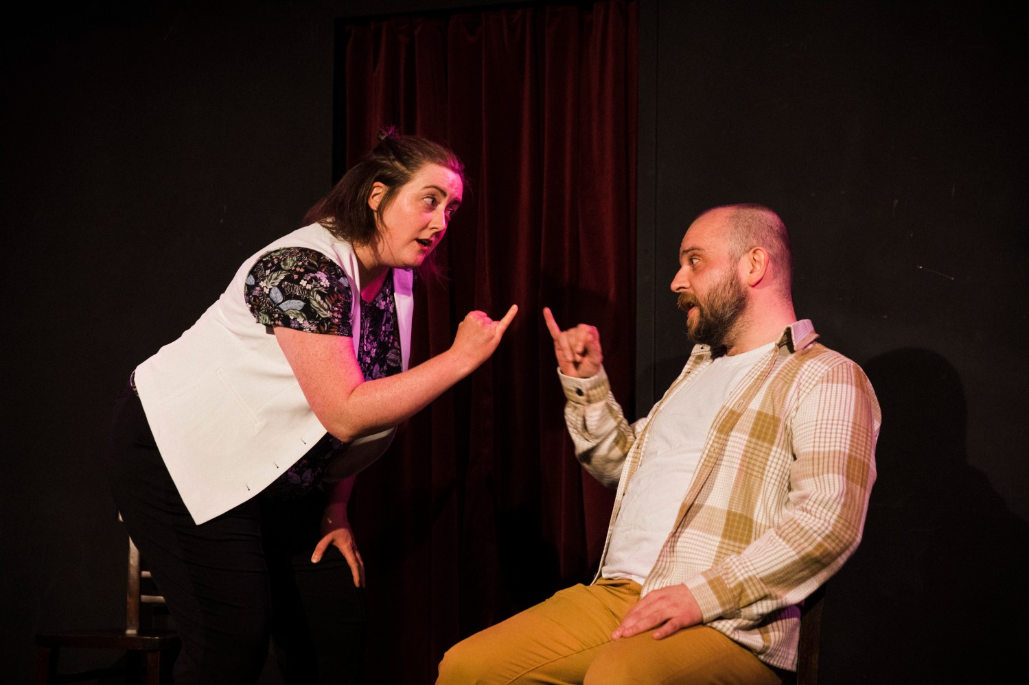 Two people energetically gesturing at each other on stage with a red curtain backdrop.