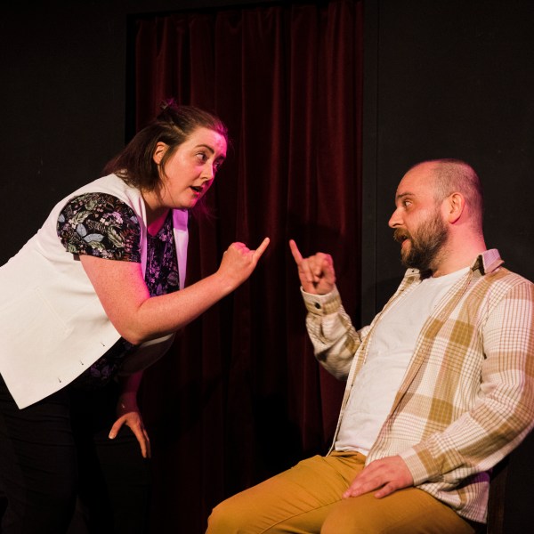 Two people energetically gesturing at each other on stage with a red curtain backdrop.