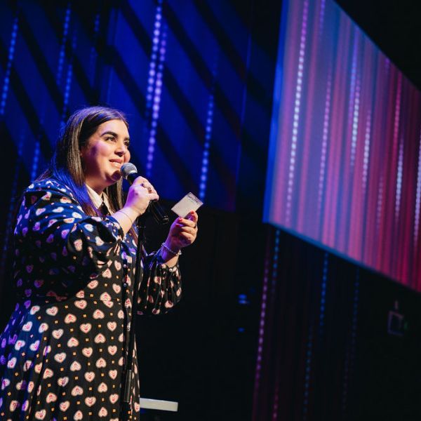 Person speaking into a microphone on stage with colorful lights.