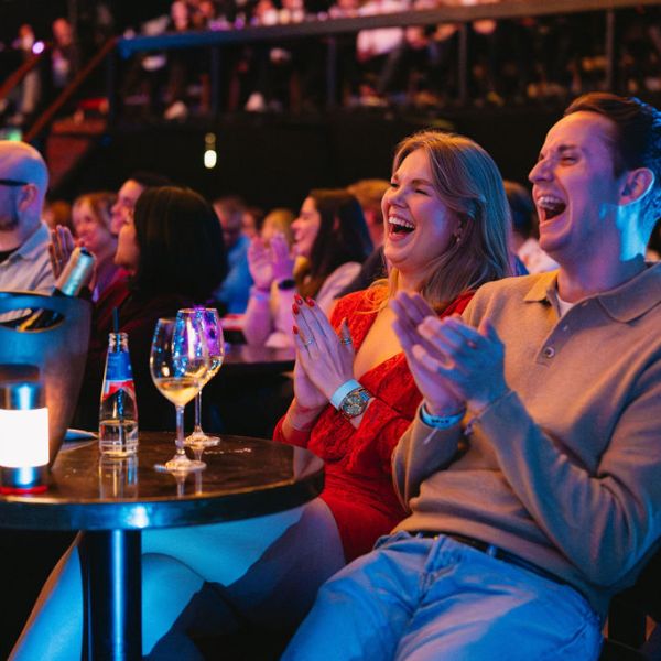 Audience clapping and smiling in a dimly lit venue with drinks on tables.