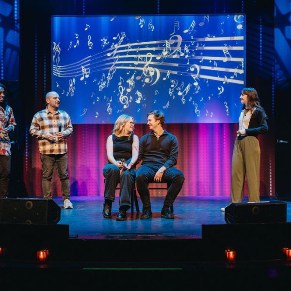 Six people on stage, two seated in center, musical notes on screen behind them, colorful lighting.