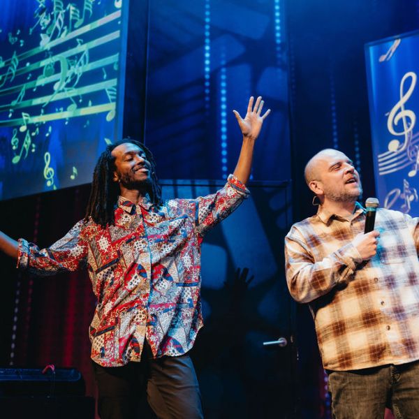 Two men on stage with microphones, colorful music notes projected behind them.
