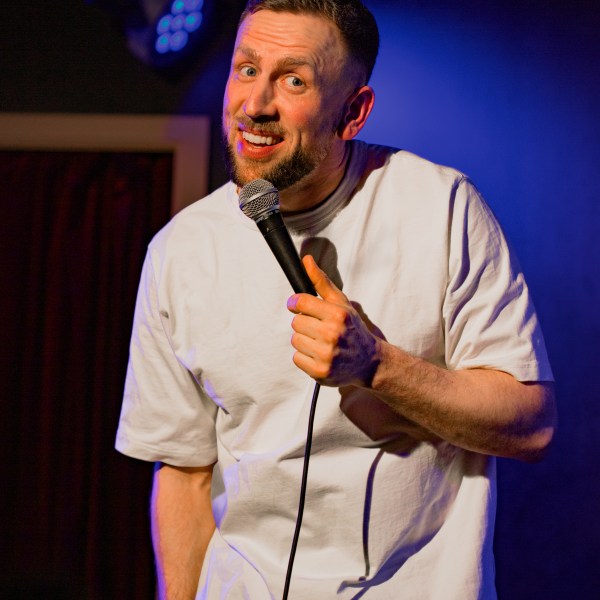 Comedian on stage in white shirt holding a microphone, with blue spotlight behind.