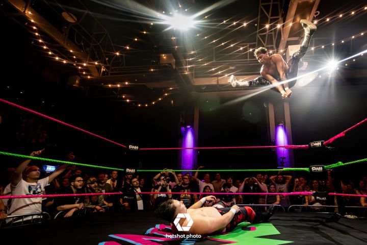 Wrestler in mid-air above opponent in a ring with a cheering crowd and bright lights.