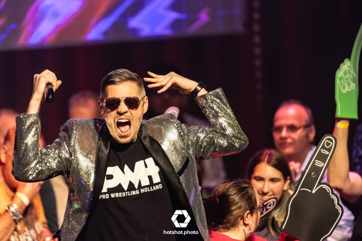 Man in sparkly jacket singing on stage with crowd and foam hands.
