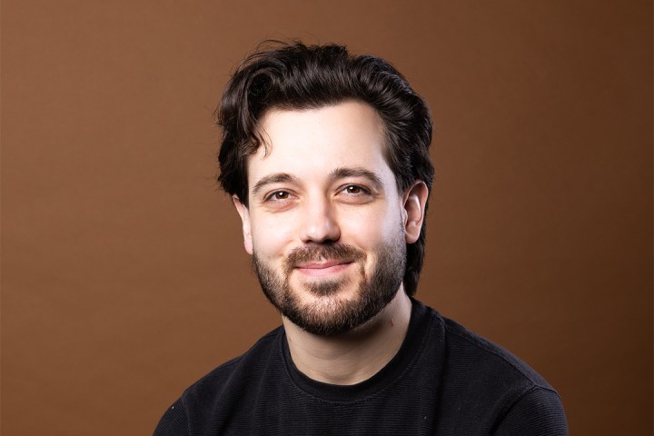 Man with a beard and dark hair smiling against a brown background.