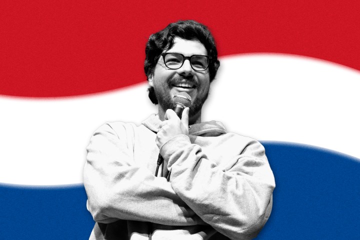 Man with glasses smiling, holding a microphone, with red, white, and blue striped background.