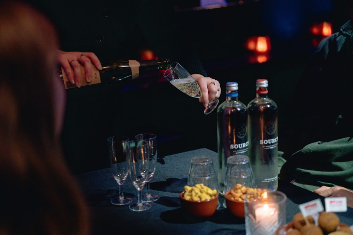 Person pouring champagne into glass beside bottles and snacks on table.
