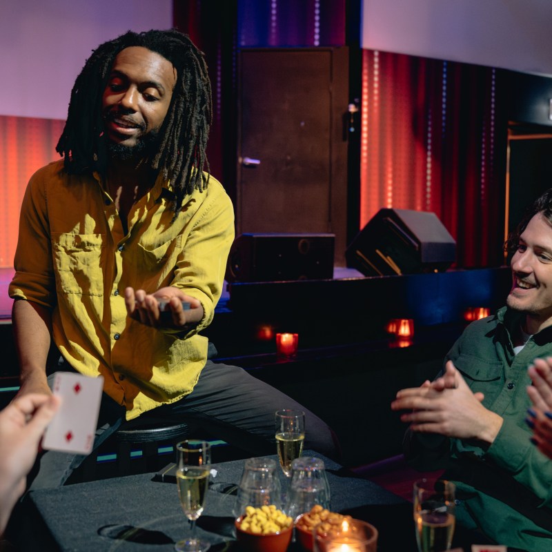 Person in yellow shirt performing card trick for seated audience in dimly lit room.