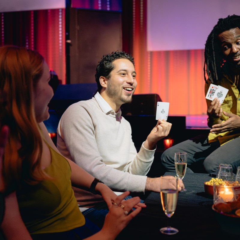 People enjoying a card trick at a table with drinks and snacks.