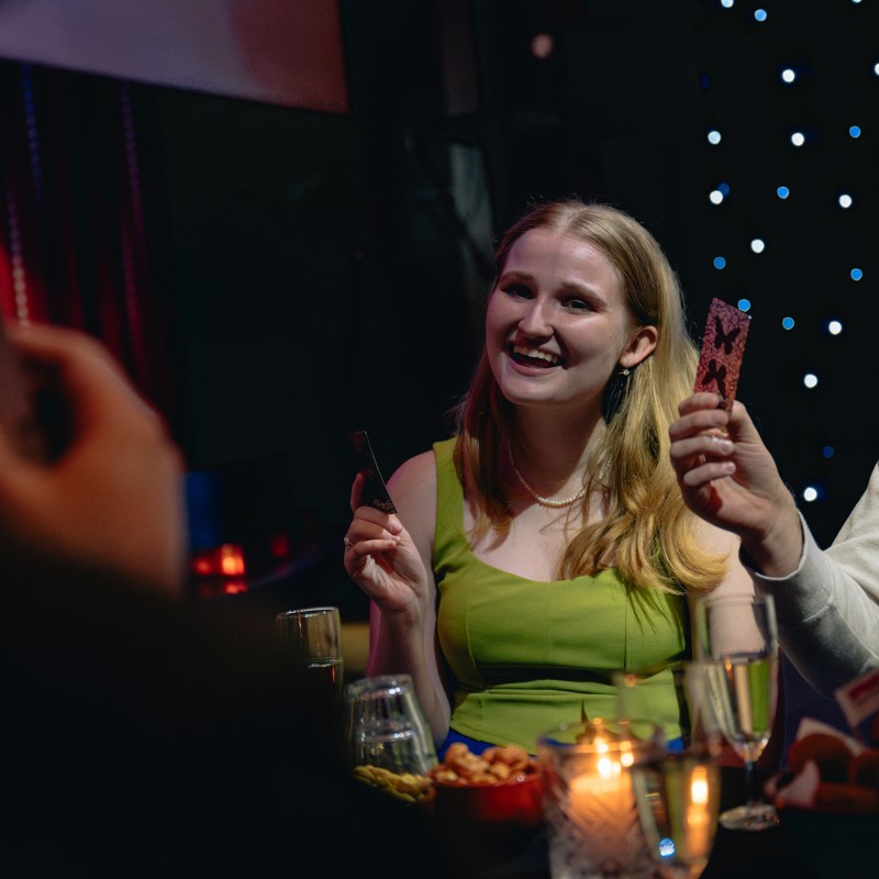People at a table playing cards, a woman smiling and holding a card, with snacks and drinks around.