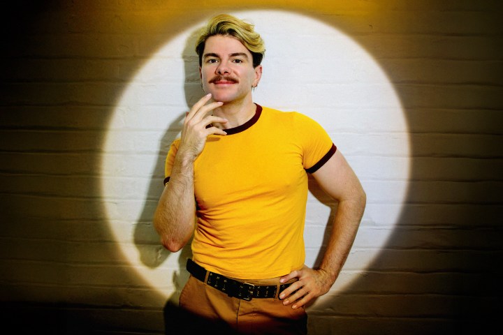 Person with mustache in yellow shirt under spotlight, posed with hand on chin against brick wall.