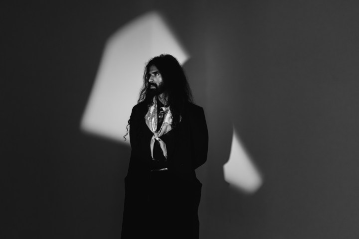 Man with long hair and beard stands in shadowy light, black and white photo.