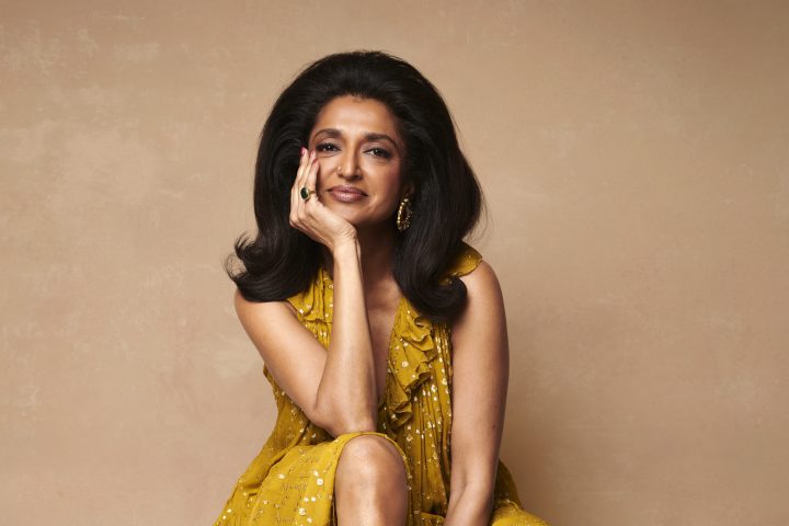 Person in a yellow dress sitting on a chair against a beige background, wearing black boots, smiling.