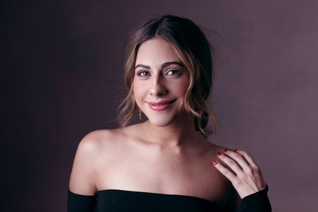 Woman with shoulder-length hair in off-shoulder black top, smiling at camera with hand near collarbone.
