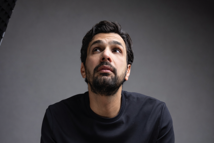 Man sitting on stool, looking upwards, in dark T-shirt and pants against a gray background.