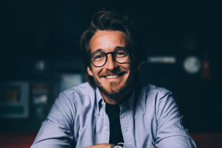 Smiling man with glasses and blue shirt sitting in red booth.
