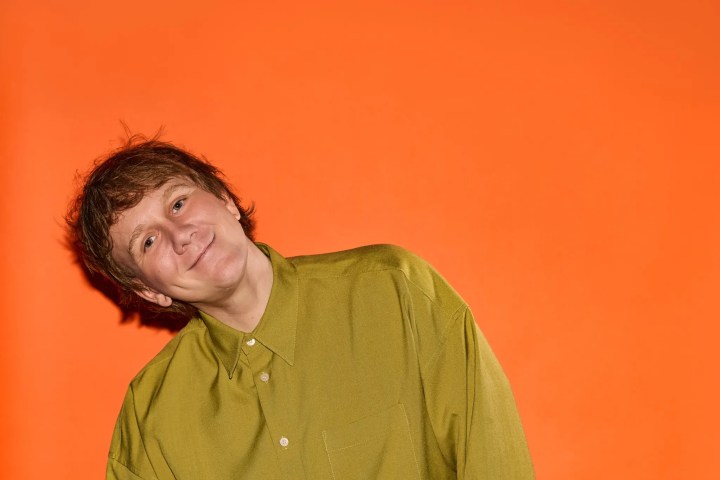 Person in green shirt leaning head to the side against an orange background.