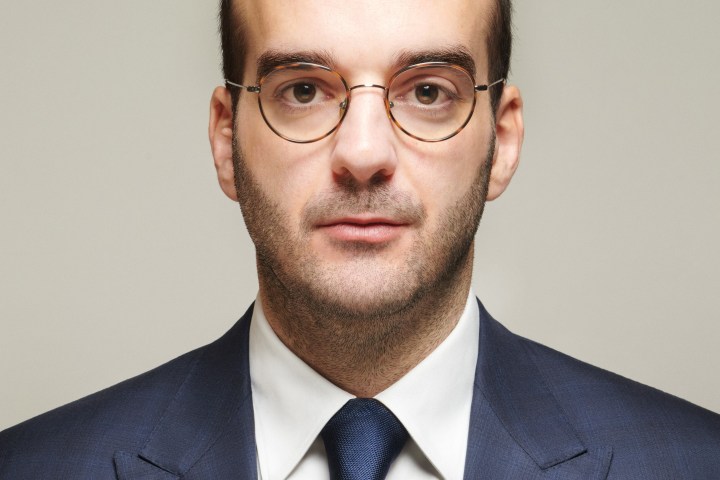 Man with short hair, glasses, wearing a blue suit and tie against a neutral background.