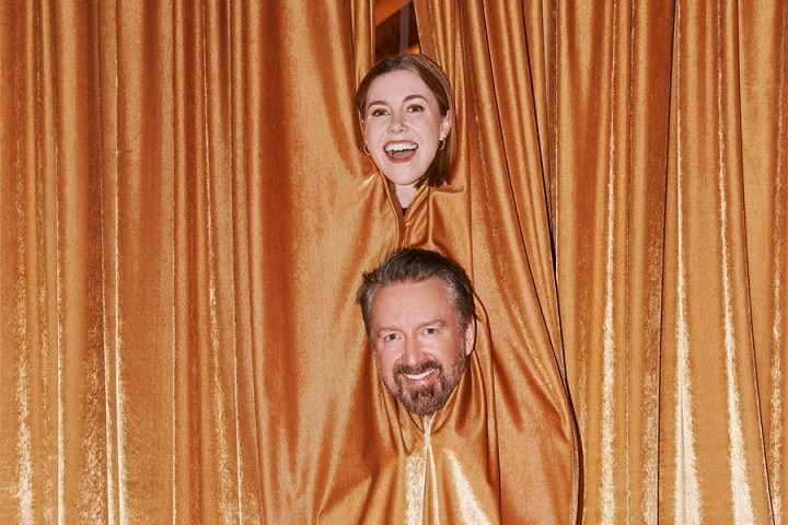 Two people smiling, peeking through golden curtains.