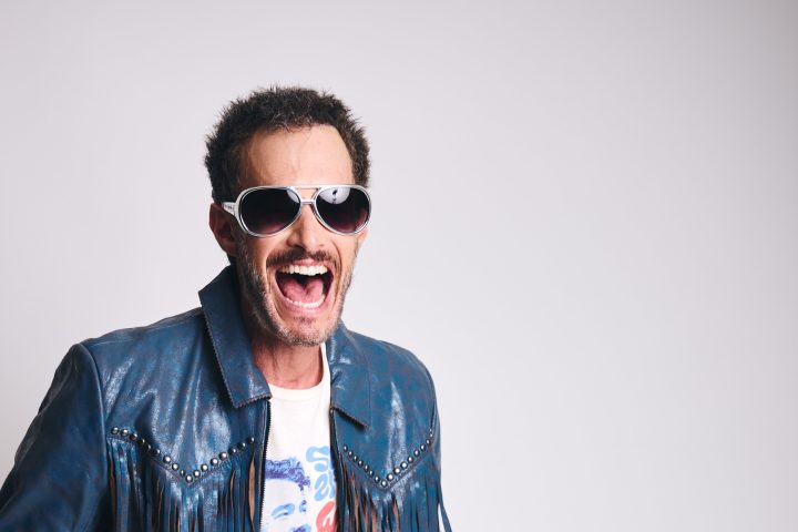 Man in blue fringe jacket and sunglasses laughing, wearing jeans.