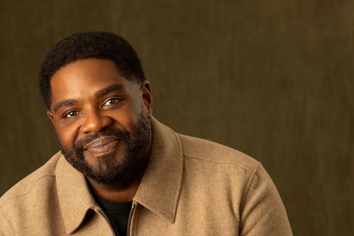 Man with short hair and beard smiling, wearing a tan jacket against a brown background.