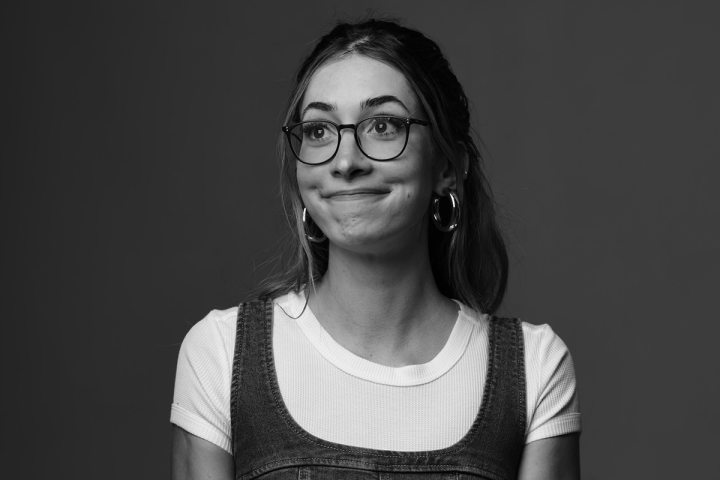 Black and white photo of a person with glasses, wearing a white shirt and denim dress, smiling slightly.