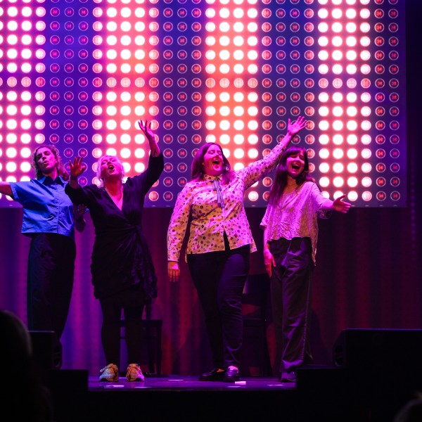 Four people on stage under colorful lights, waving with one arm raised.