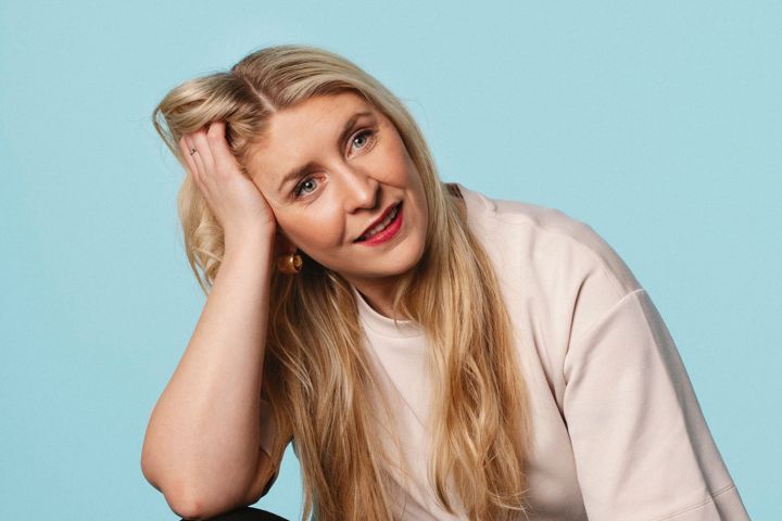 Smiling woman with long blonde hair, resting head on hand against blue background.