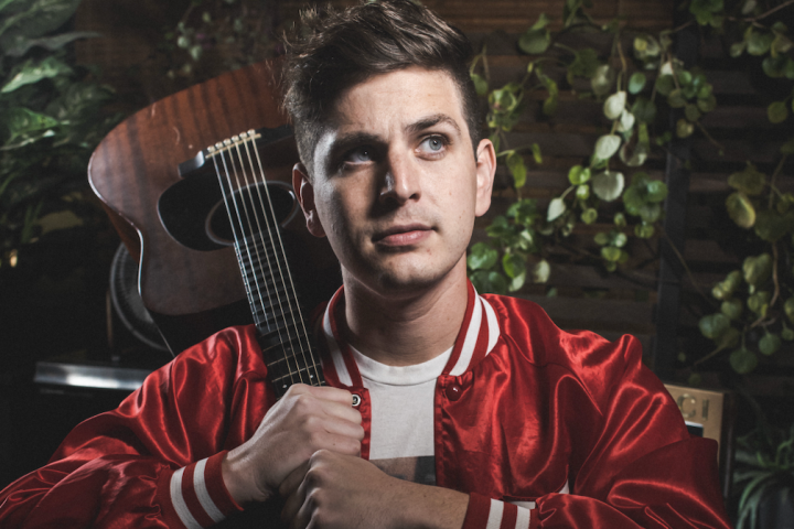 Person in red jacket holds guitar against green leafy background.