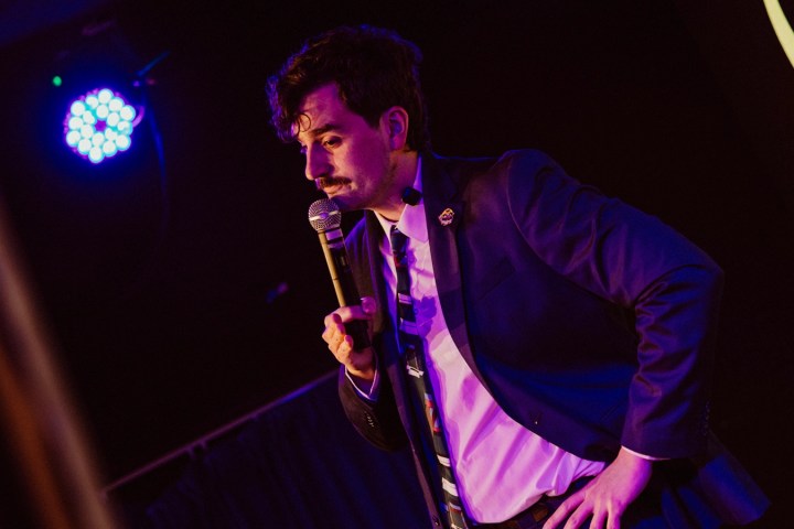 Man in suit performing stand-up comedy on stage with a microphone.