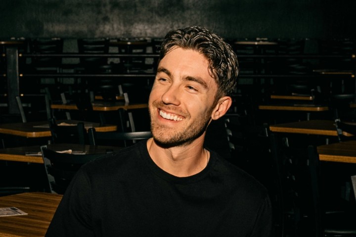 Man in black shirt smiling in dark room with empty chairs and tables.